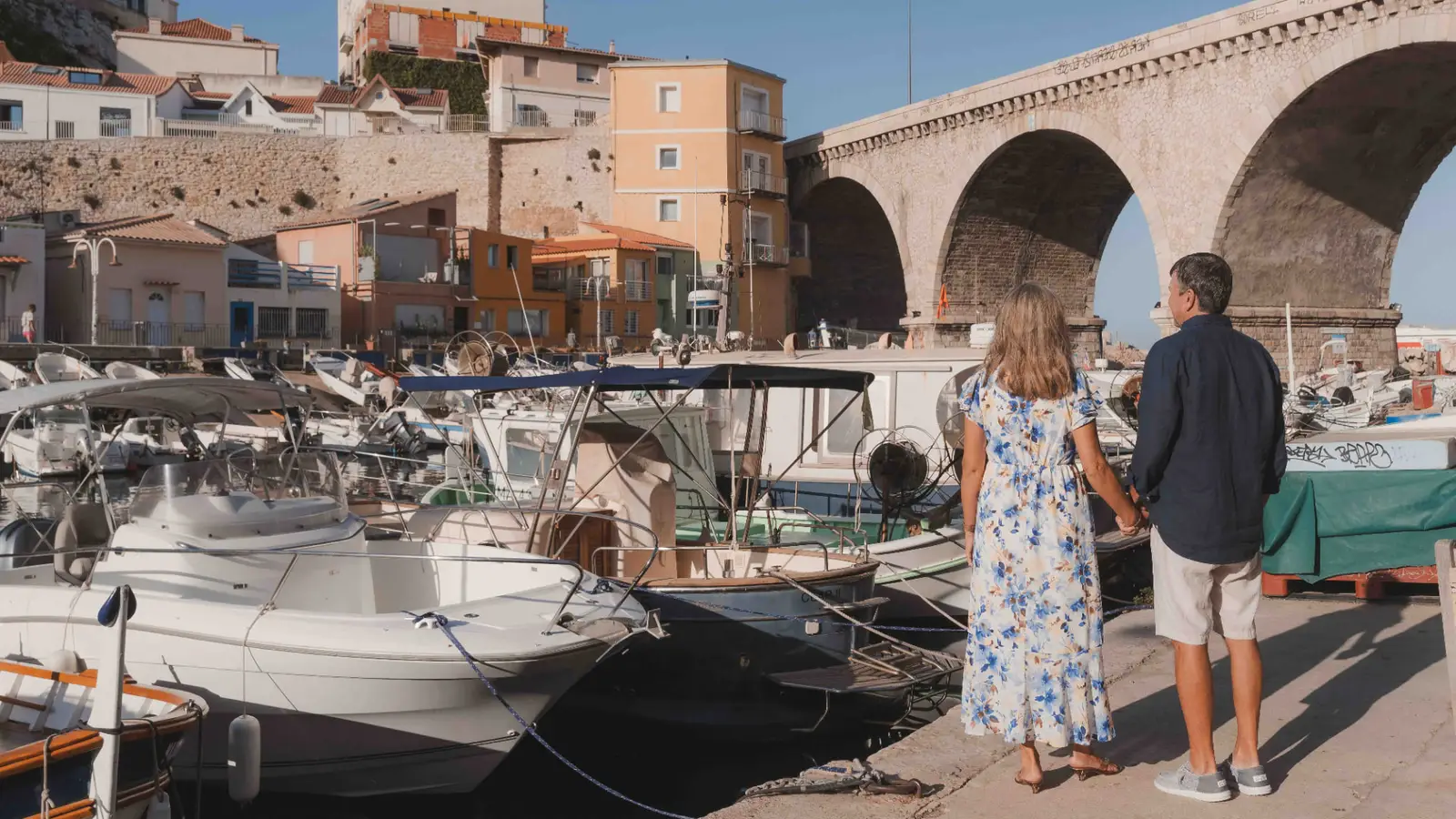 Couple holding hands in front of the moored fishing boats and stone arches of Vallon des Auffes, Marseille