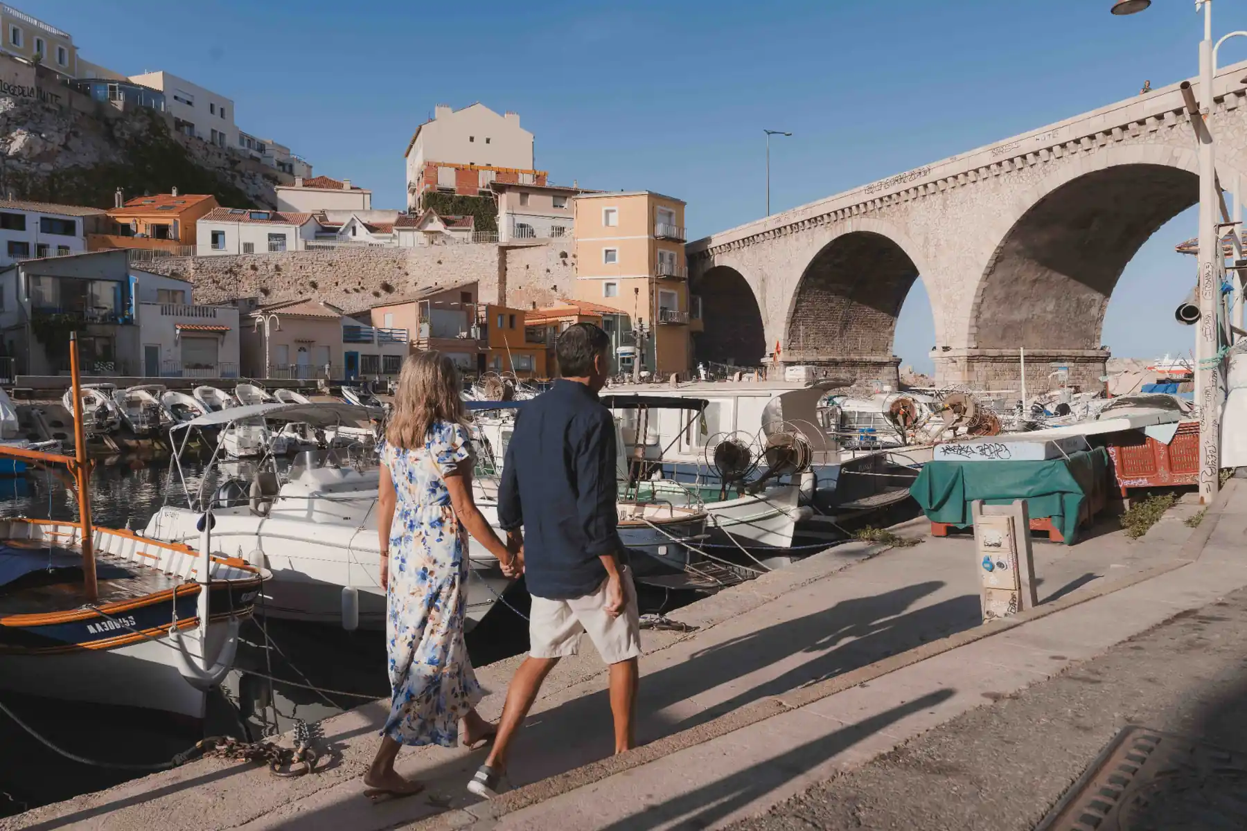 American couple walking hand in hand past the fishing boats and the stone bridge of Vallon des Auffes, Marseille